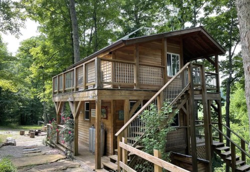A wooden two-story cabin surrounded by trees, featuring a staircase and a porch with railings.