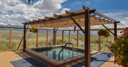 A shaded hot tub area with a wooden pergola, surrounded by greenery and mountains under a cloudy sky.