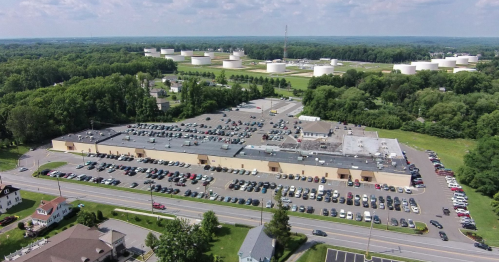 Aerial view of a large building with a parking lot, surrounded by green trees and industrial storage tanks in the background.