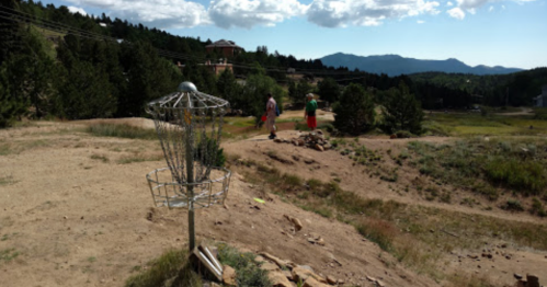 Two people walking on a dirt path in a mountainous area, with a disc golf basket in the foreground.
