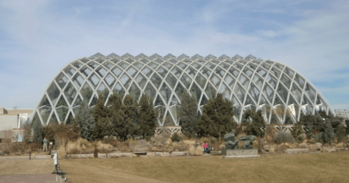 A large, modern glass greenhouse with a geometric design, surrounded by greenery and a clear blue sky.