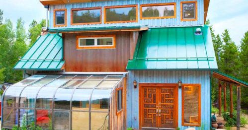 A unique blue and green house with large windows and a greenhouse attached, surrounded by trees.