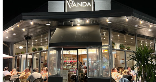 A lively restaurant at night, featuring large windows, outdoor seating, and a bright sign reading "Vanda."