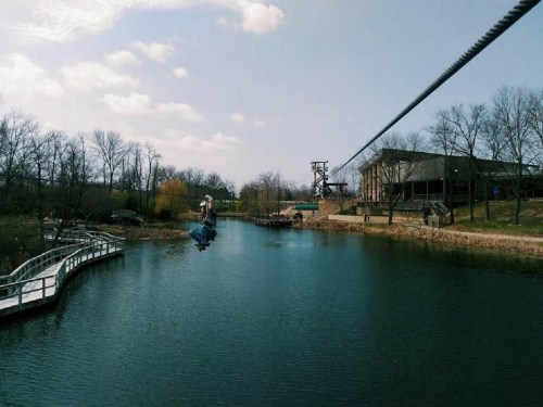 A person zip-lining over a calm lake, with trees and a building in the background under a partly cloudy sky.