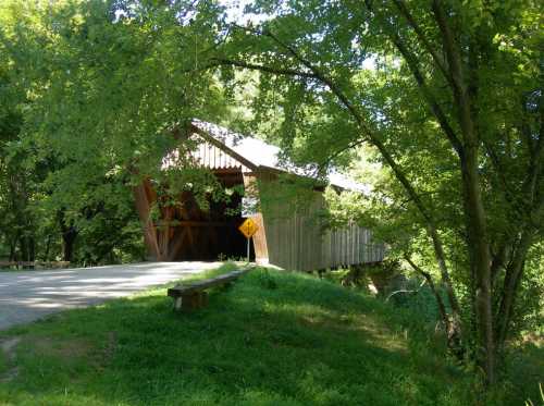 A wooden covered bridge surrounded by lush green trees and grass, with a warning sign nearby.