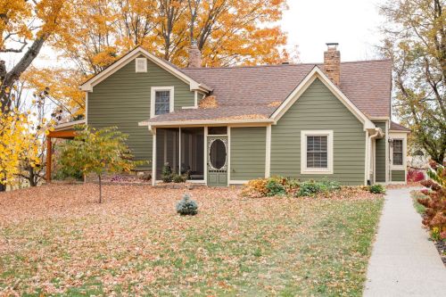 A charming green house surrounded by colorful autumn leaves and trees, with a welcoming front porch.