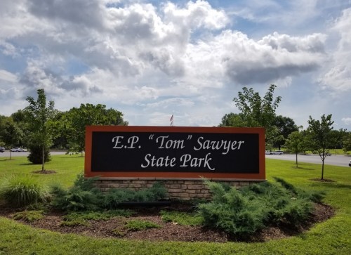 Sign for E.P. "Tom" Sawyer State Park, surrounded by greenery and under a partly cloudy sky.