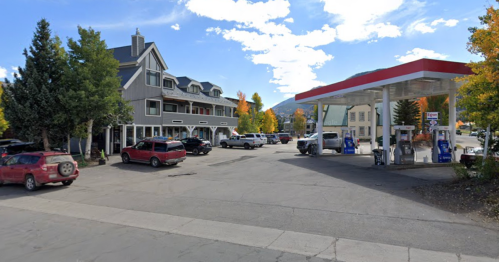 A gas station next to a multi-story building, surrounded by trees and parked cars under a blue sky with clouds.
