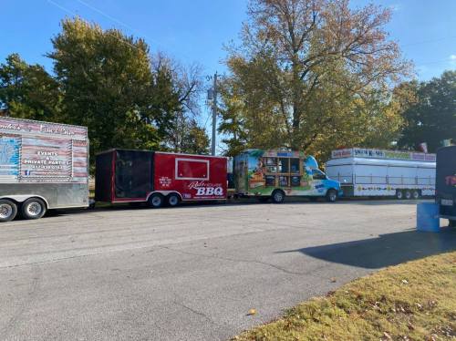 A row of food trucks and trailers parked along a tree-lined street on a sunny day.