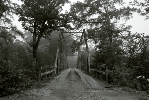 A foggy, black-and-white image of an old metal bridge surrounded by trees, leading down a gravel road.