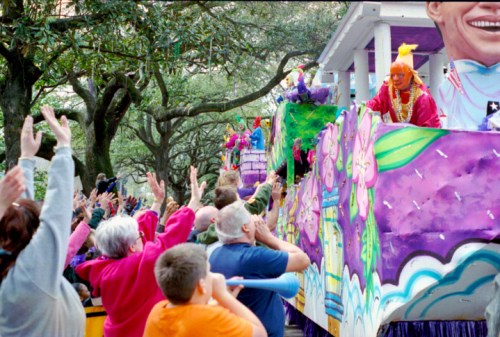 A colorful parade float with performers, surrounded by a cheering crowd under trees. Festive atmosphere.