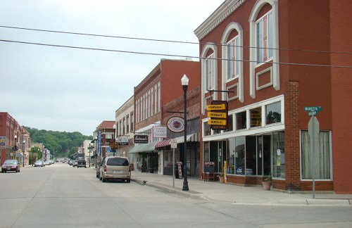 A quiet downtown street lined with brick buildings, shops, and a parked car under a cloudy sky.