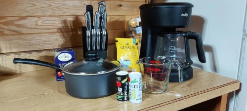 A kitchen countertop with a coffee maker, saucepan, measuring cup, salt, pepper, and a knife set against a wooden backdrop.