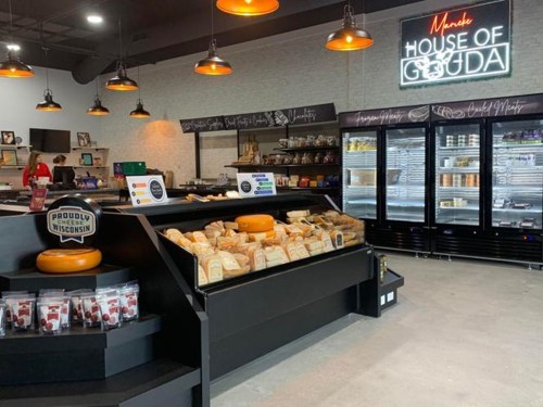 Interior of a cheese shop featuring a display of cheeses, a refrigerated section, and a sign reading "House of Gouda."