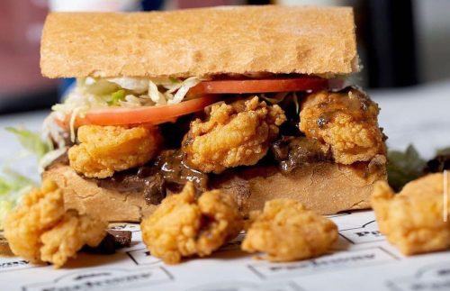 A close-up of a sandwich filled with fried shrimp, lettuce, and tomato, served on a crusty bread with fried pieces beside it.