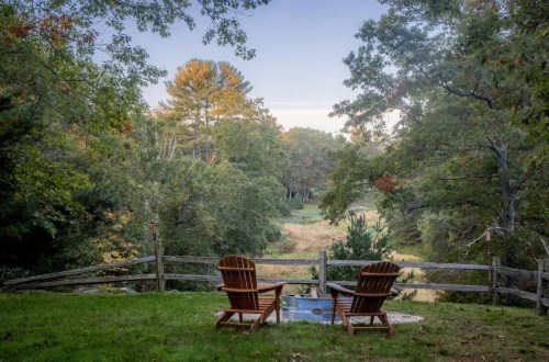 Two wooden chairs facing a serene landscape with trees and a grassy field, framed by a rustic wooden fence.