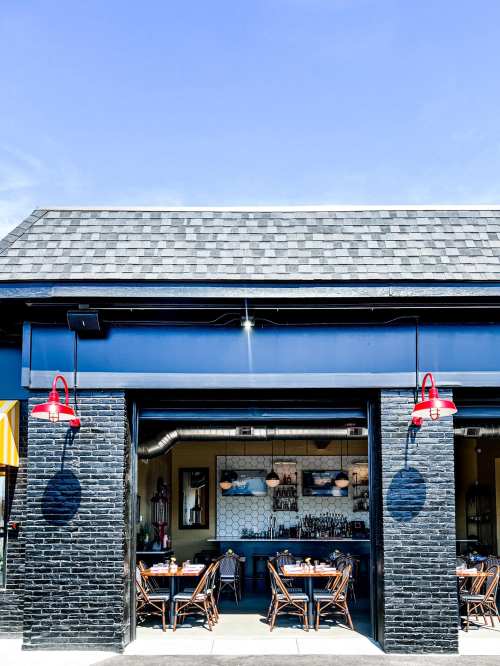 A modern restaurant exterior with open garage doors, outdoor seating, and red pendant lights against a clear blue sky.