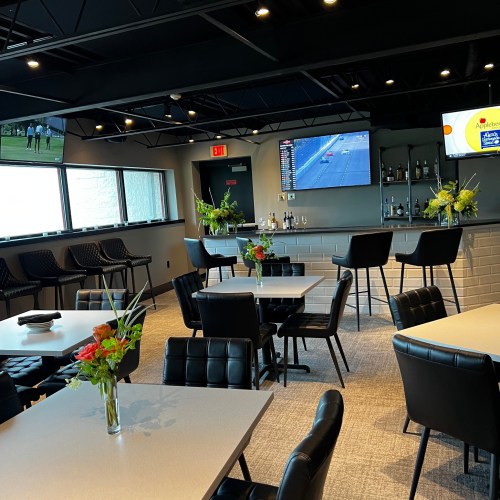 Modern restaurant interior with tables, bar area, and large TV displaying a sports event. Decorated with flowers.