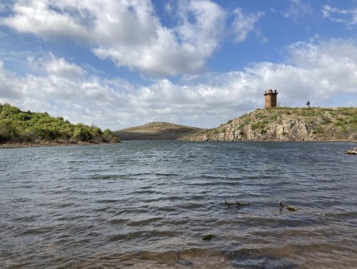 A scenic view of a lake surrounded by hills, featuring a tower on a distant hill under a partly cloudy sky.