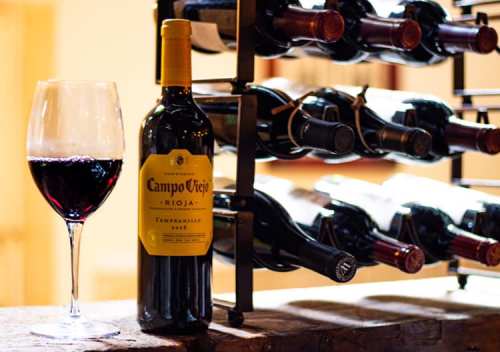 A bottle of Campo Viejo Rioja Tempranillo next to a glass of red wine, with a wine rack in the background.