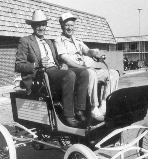 Two men in hats sit side by side in a vintage car, smiling at the camera in a sunny outdoor setting.