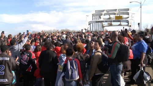A large crowd gathers on Edmund Pettus Bridge, holding signs and taking photos during a commemorative event.