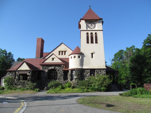 Historic stone building with a clock tower, red roof, and surrounding greenery under a clear blue sky.