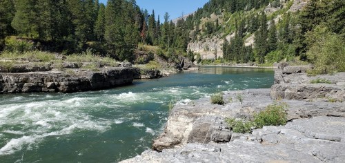 A serene river flows between rocky banks, surrounded by lush green trees and steep hills in a natural landscape.