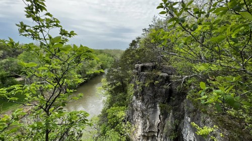 A scenic view of a river winding through lush greenery and rocky cliffs under a cloudy sky.