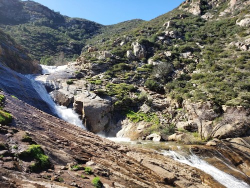 A scenic view of a waterfall cascading down rocky terrain, surrounded by lush greenery and hills under a clear blue sky.