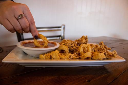 A hand dips a fried appetizer into a small bowl of sauce on a white plate, with more fried items beside it.