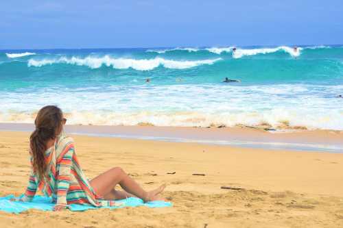 A person sits on the beach, watching surfers ride waves under a blue sky with fluffy clouds.