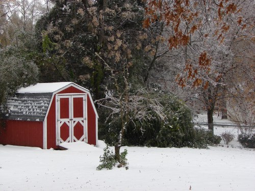 A red barn surrounded by snow and icy trees, with a white landscape and a fence in the background.