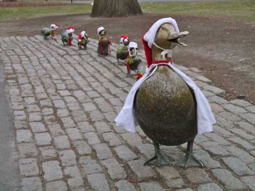 A line of bronze duck sculptures wearing Santa hats and scarves on a cobblestone path in a park.