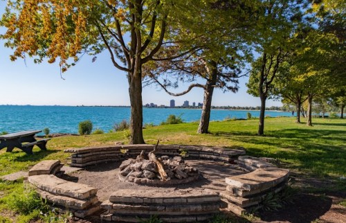 A circular stone fire pit surrounded by trees, overlooking a calm lake and city skyline in the distance.