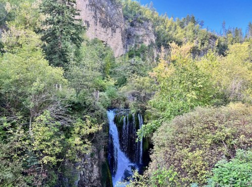 A serene waterfall cascades through lush greenery, surrounded by rocky cliffs and vibrant trees under a clear blue sky.