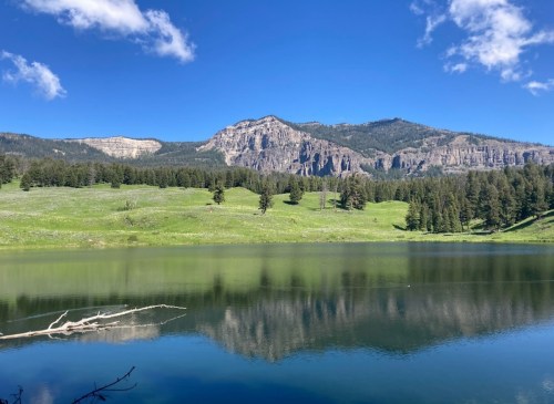 A serene lake reflects green hills and rocky mountains under a clear blue sky with scattered clouds.