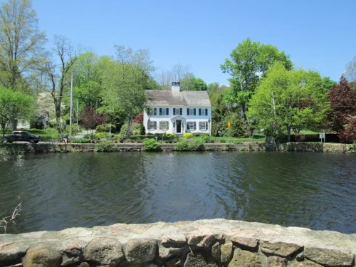A charming white house surrounded by lush greenery, reflecting in a calm lake under a clear blue sky.