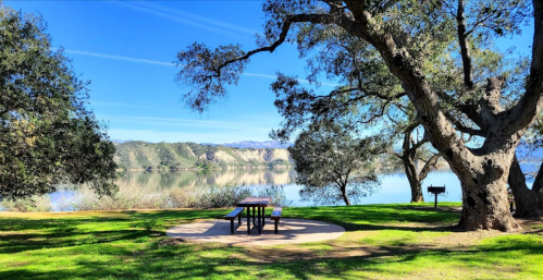 A serene lakeside scene with a picnic table, surrounded by trees and mountains reflecting in the calm water.