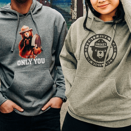 Two people wearing hoodies with Smokey Bear designs, standing outdoors with a blurred background.