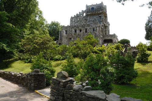 A stone castle surrounded by lush greenery and trees, set on a hillside under a clear blue sky.