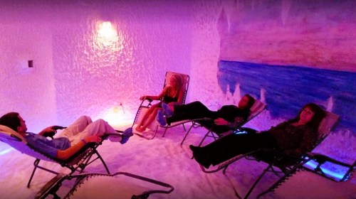 Three people relax in reclining chairs inside a salt room, surrounded by pink and blue lighting.