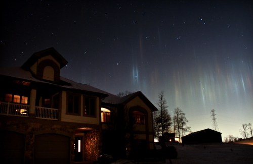A house at night under a starry sky with colorful auroras and silhouettes of trees and a barn in the background.