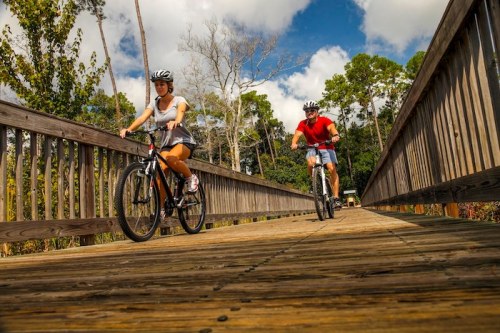 Two cyclists ride on a wooden bridge surrounded by trees under a partly cloudy sky.
