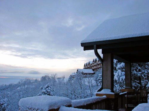 A snowy landscape with a wooden structure in the foreground and a distant building surrounded by trees.