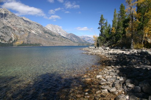 A serene lakeside view with clear water, rocky shore, and mountains under a blue sky with scattered clouds.