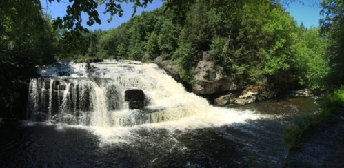 A scenic waterfall cascading over rocks, surrounded by lush green trees and a clear blue sky.