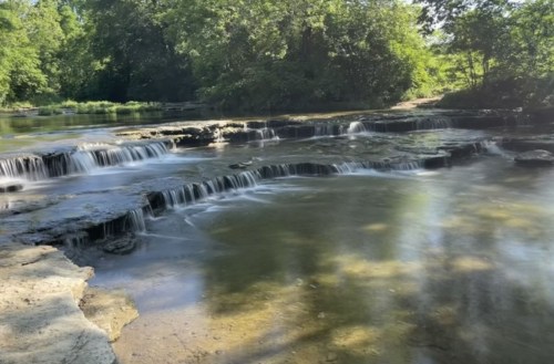 A serene river scene with gentle waterfalls cascading over rocks, surrounded by lush green trees and sunlight.