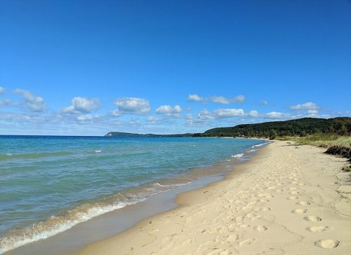 A serene beach scene with gentle waves, soft sand, and a clear blue sky dotted with clouds. Green hills in the background.