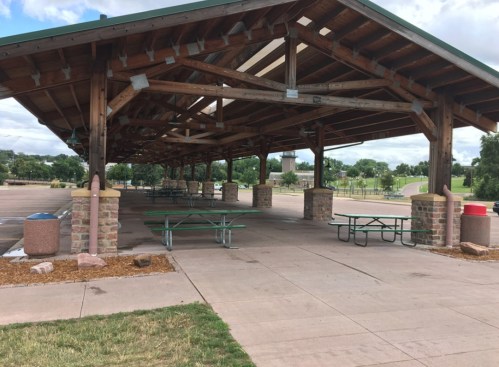 A spacious wooden pavilion with picnic tables, surrounded by grass and a paved area, under a partly cloudy sky.